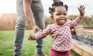 Ein Baby lernt laufen! Ein kleines Mädchen in einem rosa Shirt mit Schmetterlingen macht seine ersten Schritte im Garten, gehalten von einer erwachsenen Hand.