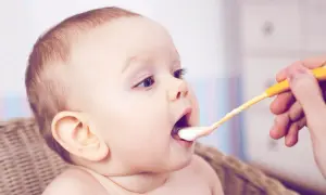 Ein Baby bekommt den ersten anfangsbrei. Die gelbe Löffelspitze mit Brei nähert sich dem offenen Mund.