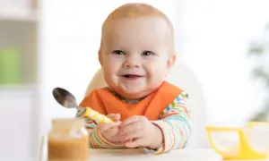 Glückliches Baby mit Lätzchen und Löffel, bereit für die Beikost. Zeichen der Freude beim Essen.