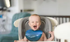 Ein glückliches Baby mit einem blauen Lätzchen sitzt im Hochstuhl und freut sich über seinen ersten Brei.