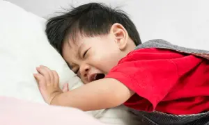Ein weinendes Baby haut sich auf den Kopf, liegt auf einem Kissen und trägt ein rotes Oberteil.