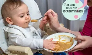 Ein Baby sitzt im Hochstuhl und bekommt Brei gefüttert. Die Hände des Babys sind voller Essen. Tipps für Baby essen von Expert:innen.