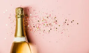 Goldene Sektflasche mit bunten Streuseln und Sternen auf rosa Hintergrund. Ein Glas Sekt stillen für den besonderen Moment.