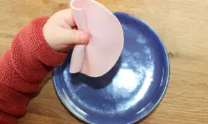Kleines Baby mit rotem Pullover hält Wurstscheibe über blauen Teller. Ab wann darf Baby Wurst essen?