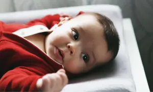 Ein süßes Baby in rotem Strampler liegt auf einer Wickelauflage. Es schaut neugierig in die Kamera, als ob es gerade vom Wickeltisch gefallen wäre und sich wundert, wie es hierher kam.