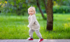 Ein fröhliches, blondes Kind, circa 2 Jahre alt, läuft auf einem Weg im Park. Es trägt einen Strickcardigan und rote Turnschuhe.