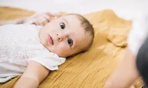 Ein neugieriges Baby, etwa 3 Monate alt, liegt auf einer senfgelben Decke und blickt mit großen Augen in die Kamera.