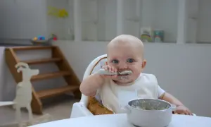 Kleines Baby mit blauen Augen isst begeistert Baby Naturjoghurt. Löffel und Lätzchen sind voller leckerer Reste.
