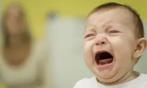 Ein Baby kreischt schrill, die Augen geschlossen, der Mund weit geöffnet. Im Hintergrund eine unscharfe Person.
