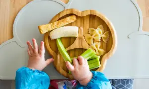 Baby obst ab wann? Ein Kleinkind greift nach Banane, Gurke und Nudeln auf einem Bambusteller.