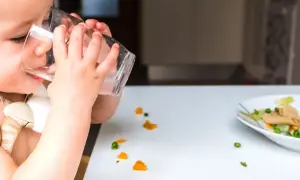 Kleines Kind trinkt aus Glas, daneben ein Teller mit Brei und verstreuten Gemüsestückchen. Wie viel Wasser bei Beikost?
