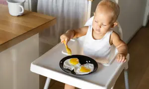 Kleines Kind im Hochstuhl versucht, Spiegeleier mit einer Gabel zu essen. Manche Eltern sehen dies als verbotene Lebensmittel im 2. Lebensjahr.