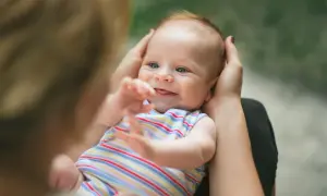 Ein lächelndes neugeborenes Baby mit blauen Augen wird von Händen gehalten. Es trägt ein gestreiftes Oberteil und streckt seine Arme aus.