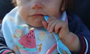Ein Baby mit Lätzchen isst mit einem blauen Löffel. Das Lätzchen zeigt Früchte und das Wort "Fruchtchen". Das ist ein Beispiel für Beikost.