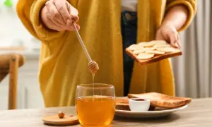 Goldener Honig tropft von einem Holzstäbchen auf ein Glas. Eine Person bereitet Toast mit Bananen und Honig zu, ein süßer Genuss beim Stillen.