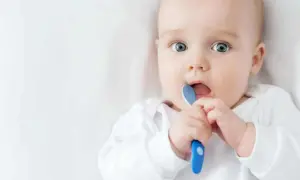 Ein Baby mit großen blauen Augen hält einen blauen Löffel und steckt ihn in den Mund. Es ist ein süßer sprung 5 baby.