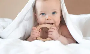 Blauäugiges Baby unter weißer Decke, kaut auf Holzring. Ab wann sabbern Babys? Oft schon vor dem Zahnen.
