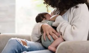 Mutter stillt unruhiges Baby, während sie auf einem Sofa sitzt. Ein Kaffee steht vielleicht in Reichweite.