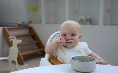 Kleines Baby mit blauen Augen isst begeistert Baby Naturjoghurt. Löffel und Lätzchen sind voller leckerer Reste.