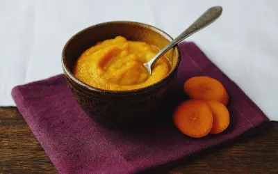 Ein Schälchen mit orangefarbenem Babybrei, daneben Karottenscheiben. Perfekt für den ersten Fisch Babybrei.