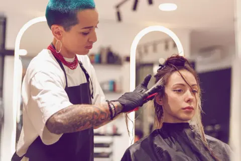 Friseurin mit blauen Haaren blondiert die Haare einer Kundin. Die Kundin wartet geduldig auf das Ergebnis, während die Friseurin sorgfältig arbeitet.