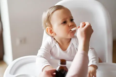 Ein Baby im Hochstuhl erhält Medikamente per Löffel. Die Eltern wechseln zwischen Ibuprofen und Paracetamol alle 4 Stunden, um Fieber zu senken.