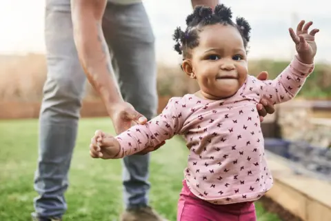 Ein Baby lernt laufen! Ein kleines Mädchen in einem rosa Shirt mit Schmetterlingen macht seine ersten Schritte im Garten, gehalten von einer erwachsenen Hand.