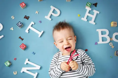Lachendes Baby mit Brille inmitten von Buchstaben. Bald beginnt die spannende Reise, ab wann sprechen Kinder?