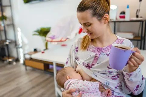 Mutter stillt Baby, trinkt Kaffee. Mögliche baby koffein symptome beachten.