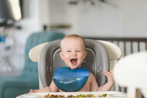 Ein glückliches Baby mit einem blauen Lätzchen sitzt im Hochstuhl und freut sich über seinen ersten Brei.