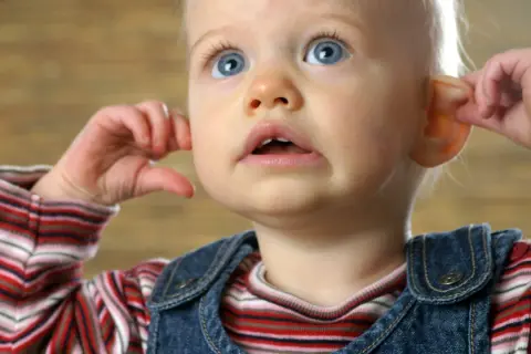 Kleines Baby kratzt sich am Ohr, die Augen weit aufgerissen.