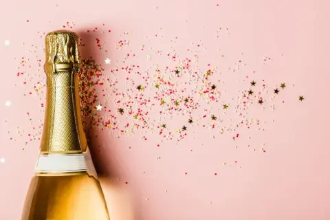 Goldene Sektflasche mit bunten Streuseln und Sternen auf rosa Hintergrund. Ein Glas Sekt stillen für den besonderen Moment.