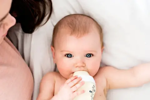 Ein Baby trinkt aus einer Flasche, während eine Person daneben liegt. Die Frage, wie lange braucht Essen bis es beim Baby ankommt, beschäftigt viele Eltern.