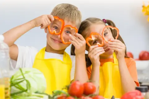Zwei Kinder mit Paprikaringen vor den Augen, die Spaß beim Kochen haben. Ein wichtiger Aspekt für gesunde Entwicklung ist der richtige kalorienbedarf kind 10 jahre.