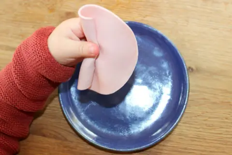 Kleines Baby mit rotem Pullover hält Wurstscheibe über blauen Teller. Ab wann darf Baby Wurst essen?