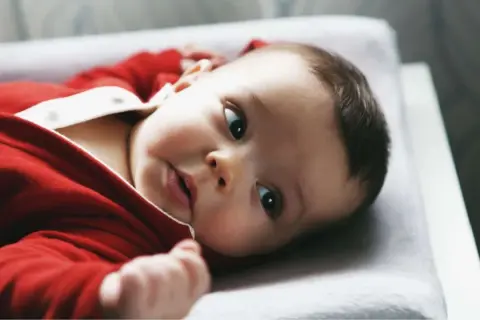 Ein süßes Baby in rotem Strampler liegt auf einer Wickelauflage. Es schaut neugierig in die Kamera, als ob es gerade vom Wickeltisch gefallen wäre und sich wundert, wie es hierher kam.