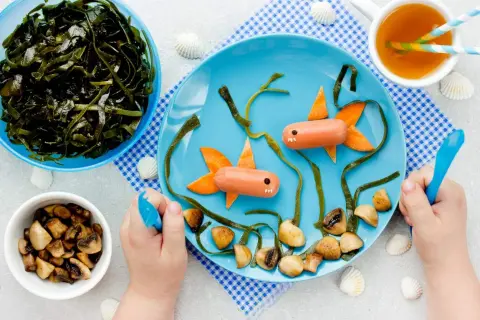 Kreativer Fischbrei für Babys: Würstchen-Fische schwimmen im Meer aus Algen und Pilzen auf einem blauen Teller.