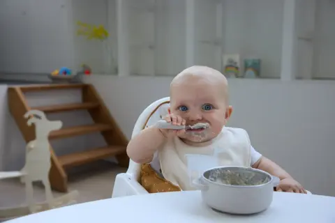 Kleines Baby mit blauen Augen isst begeistert Baby Naturjoghurt. Löffel und Lätzchen sind voller leckerer Reste.