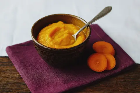 Ein Schälchen mit orangefarbenem Babybrei, daneben Karottenscheiben. Perfekt für den ersten Fisch Babybrei.