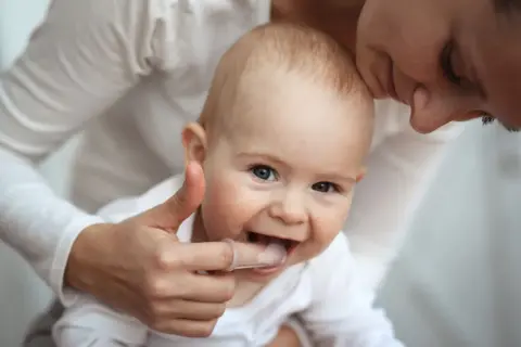 Ein Baby mit einem Daumen, der sanft die Zähne berührt, um die Zahnen Symptome zu lindern. Die Mutter küsst das Kind liebevoll.