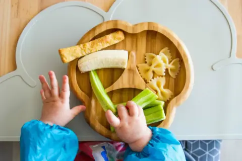 Baby obst ab wann? Ein Kleinkind greift nach Banane, Gurke und Nudeln auf einem Bambusteller.