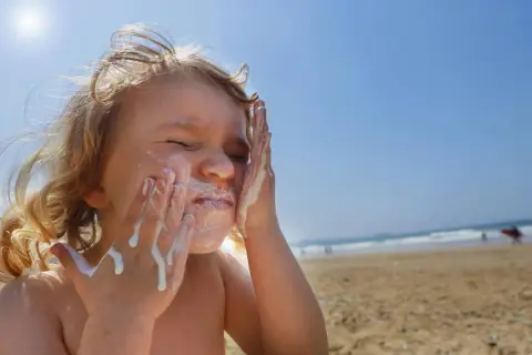 Kind schmiert sich Sonnencreme ins Gesicht. Bei Sonnenbrand helfen Hausmittel wie Quark oder Aloe Vera.