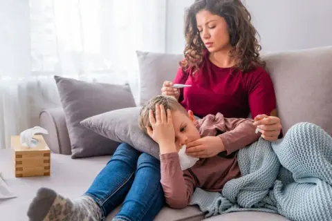 Besorgte Mutter misst Fieber bei ihrem Kind, das unter Untertemperatur leidet und schwitzt. Ein Taschentuch liegt bereit.