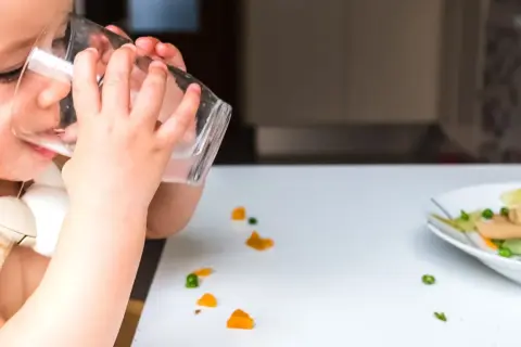 Kleines Kind trinkt aus Glas, daneben ein Teller mit Brei und verstreuten Gemüsestückchen. Wie viel Wasser bei Beikost?