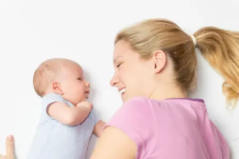 Ein lächelndes Baby, 2 Wochen alt, schaut seine Mutter an. Sie liegt neben ihm und lächelt zurück.