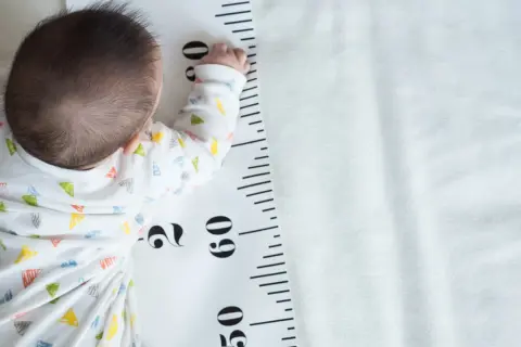 Baby liegt auf einer Messlatte, die als **Kleinwuchs Tabelle** dient. Die Messlatte zeigt Zentimeterangaben.