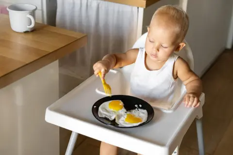 Kleines Kind im Hochstuhl versucht, Spiegeleier mit einer Gabel zu essen. Manche Eltern sehen dies als verbotene Lebensmittel im 2. Lebensjahr.