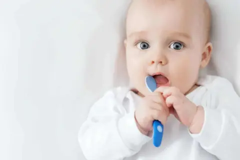 Ein Baby mit großen blauen Augen hält einen blauen Löffel und steckt ihn in den Mund. Es ist ein süßer sprung 5 baby.