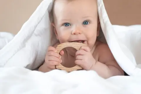 Blauäugiges Baby unter weißer Decke, kaut auf Holzring. Ab wann sabbern Babys? Oft schon vor dem Zahnen.