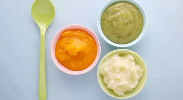 Drei Schälchen mit Babynahrung: orangefarben, grün und weiß. Welcher Joghurt Baby ist wohl am leckersten? Ein grüner Löffel liegt daneben.